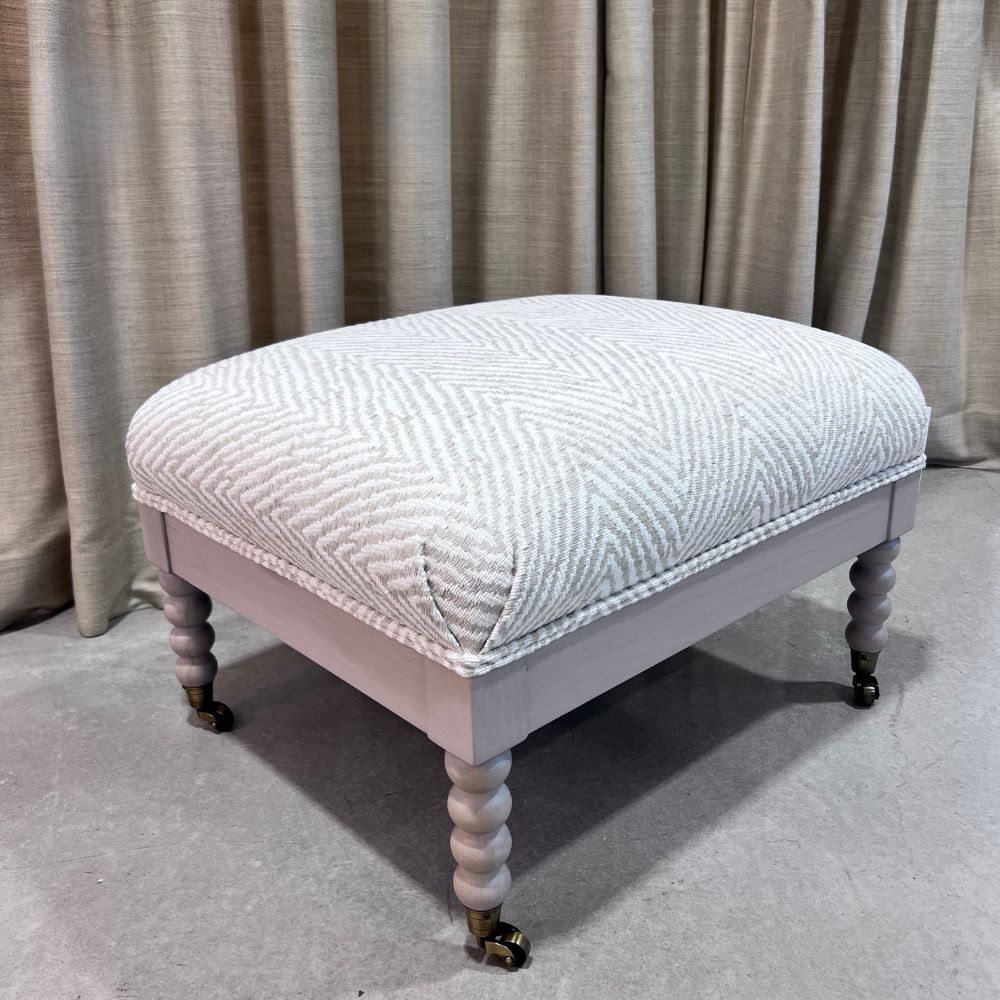 Decorative ottoman with quilted top and turned legs against a neutral curtain background. Keystone Ottoman by District Home 